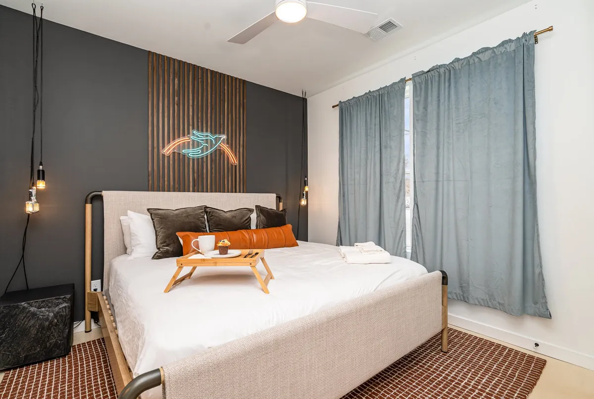 A cozy bedroom with a bed adorned with decorative pillows, a neon light feature above the headboard, and curtains softly filtering natural light. The ambiance is inviting and restful.
