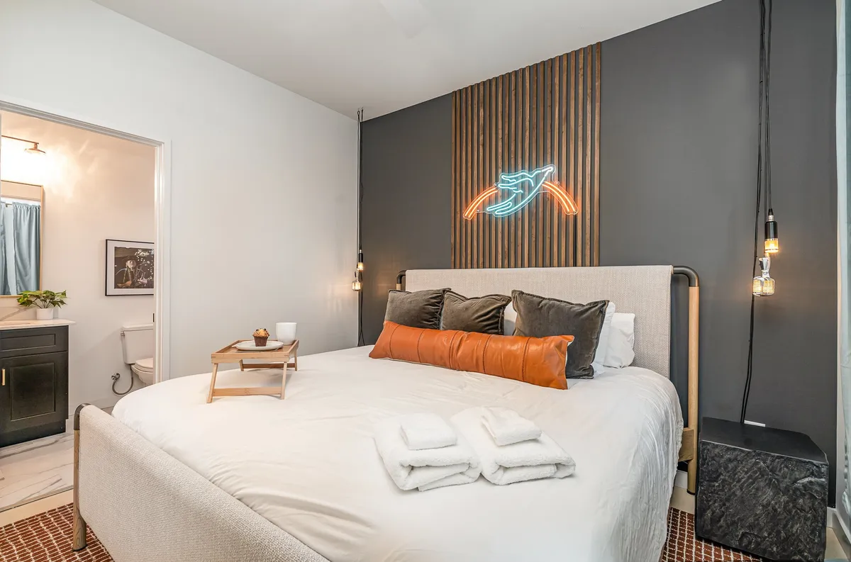 This image captures a cozy bedroom with a bed adorned with decorative pillows, a neon wall art feature, a wooden accent wall, and a nightstand with a softly glowing lamp. Soft natural light filters through the curtains, creating an inviting, restful atmosphere.
