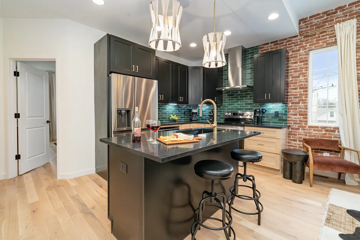 A contemporary open-concept kitchen and living area featuring a large island countertop with bar stools, modern pendant lighting, a striking brick accent wall, and stainless steel appliances. The space is inviting and clearly designed for both cooking and social gatherings, emphasizing a seamless flow between the kitchen and living space.