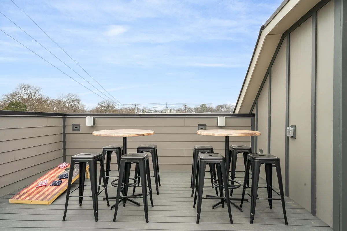 A spacious rooftop patio with wooden bar tables, black metal stools, and an inviting cornhole game set. The sky is clear, and the scene is primed for relaxation or friendly competition, with ample seating for socializing and enjoying the outdoors. The setting captures the essence of a private urban rooftop with unobstructed views, perfect for gatherings.
