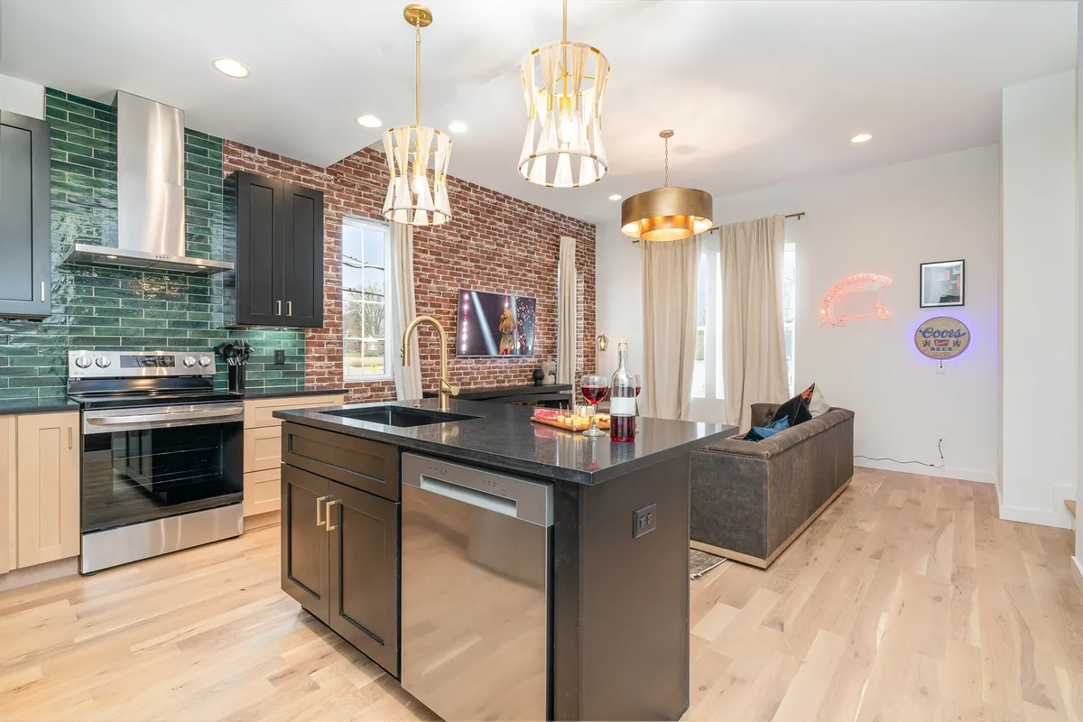 A stylish, integrated living and kitchen space with an island, exposed brick wall, chic lighting fixtures, and a sofa accented with colorful pillows. The design encourages both cooking and entertaining, with a clear visual connection between lounging and dining zones.