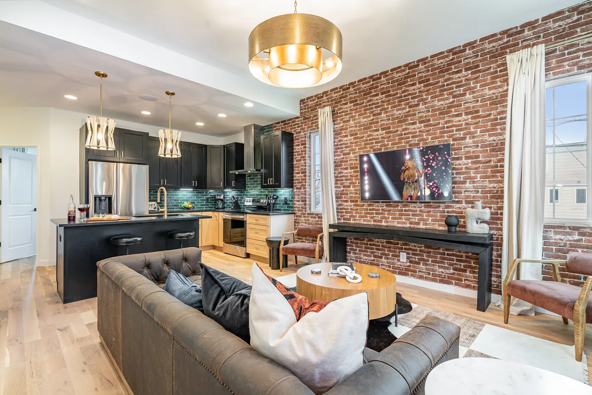 The hero image captures a spacious, modern living area featuring an exposed brick wall, a sleek and fully equipped kitchen, a plush sofa, artistic lighting, and a television displaying a live performance. The scene exudes a contemporary, lively vibe perfect for socializing or unwinding after a day in Nashville.